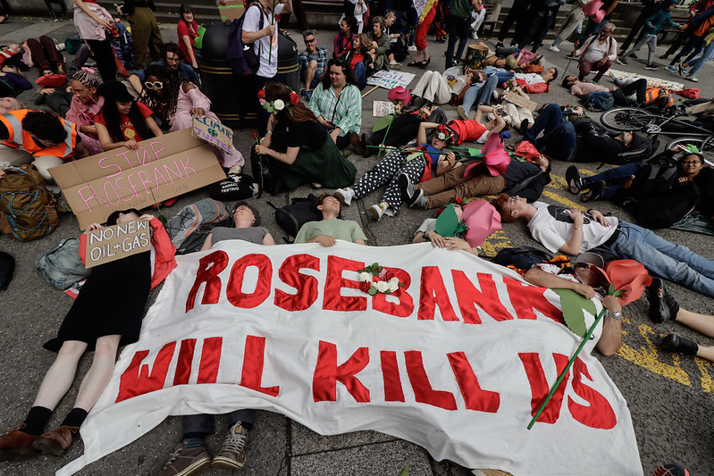 large white banner with words Rosebank will kill us in red on ground surrounded by people mostly lying round it
